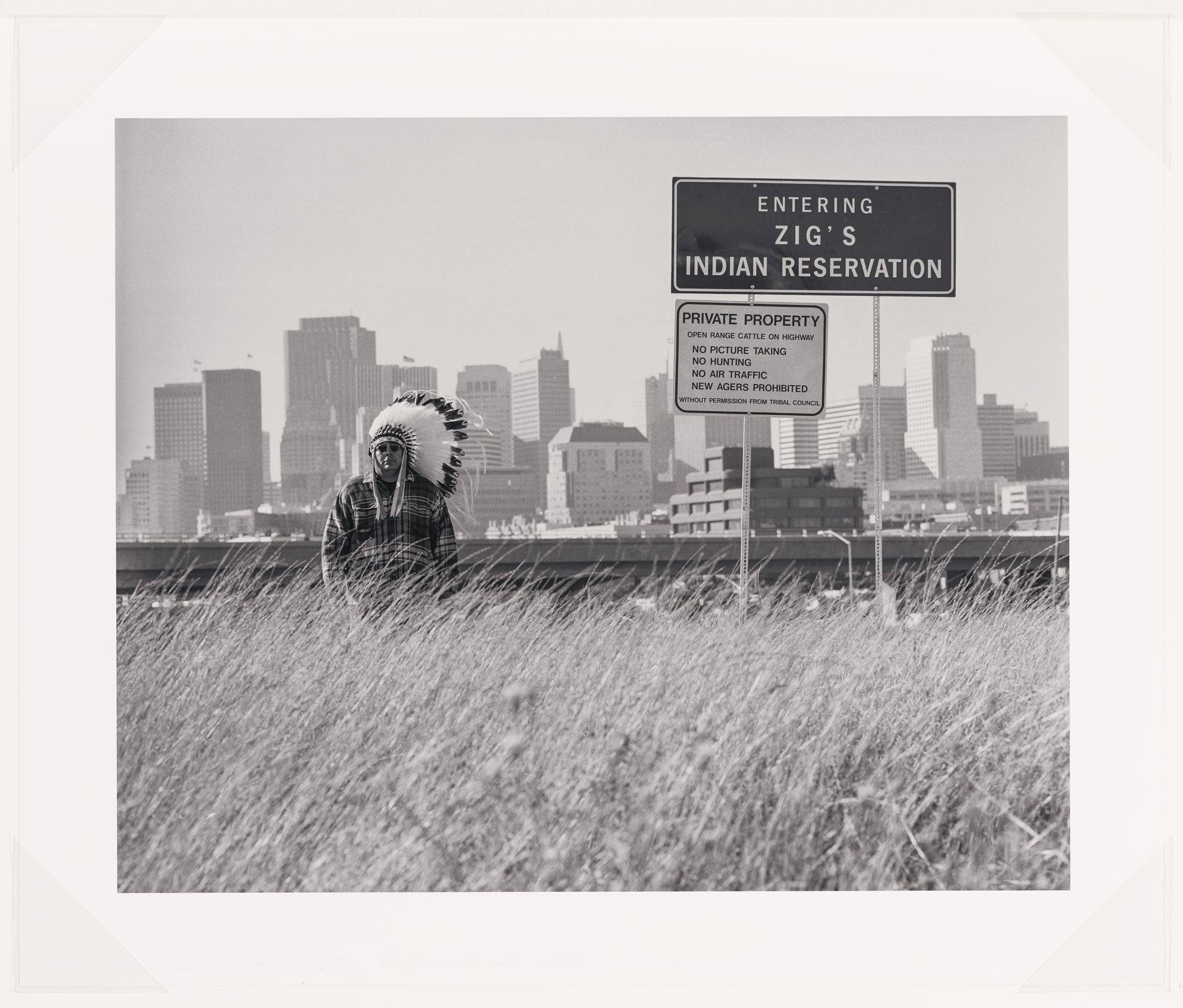 A black and white photograph that shows Zig Jackson, the artist, in a half-length portrait, standing, facing front and wearing a headdress, sunglasses, and a plaid shirt. Zig stands next to a large sign in the style of a city or landmark sign with a dark background and white lettering that states "Entering Zig’s Indian Reservation." Below is a smaller white sign with black lettering that states: "Private Property, open range cattle on highway, No Picture Taking, No Hunting, No Air Traffic, New Agers Prohibited, without permission from Tribal Council.” Zig stands in tall grass with a highway and a cityscape of San Francisco, California behind him.